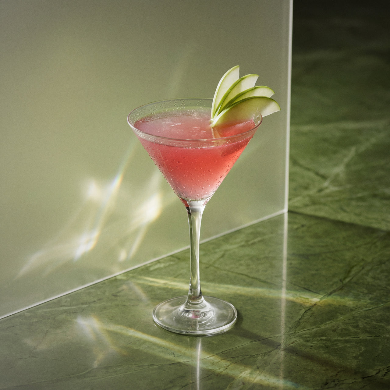 adams apple cocktail with sour apple liqueur and genever in a martini glass with a green apple slice on a reflective surface.