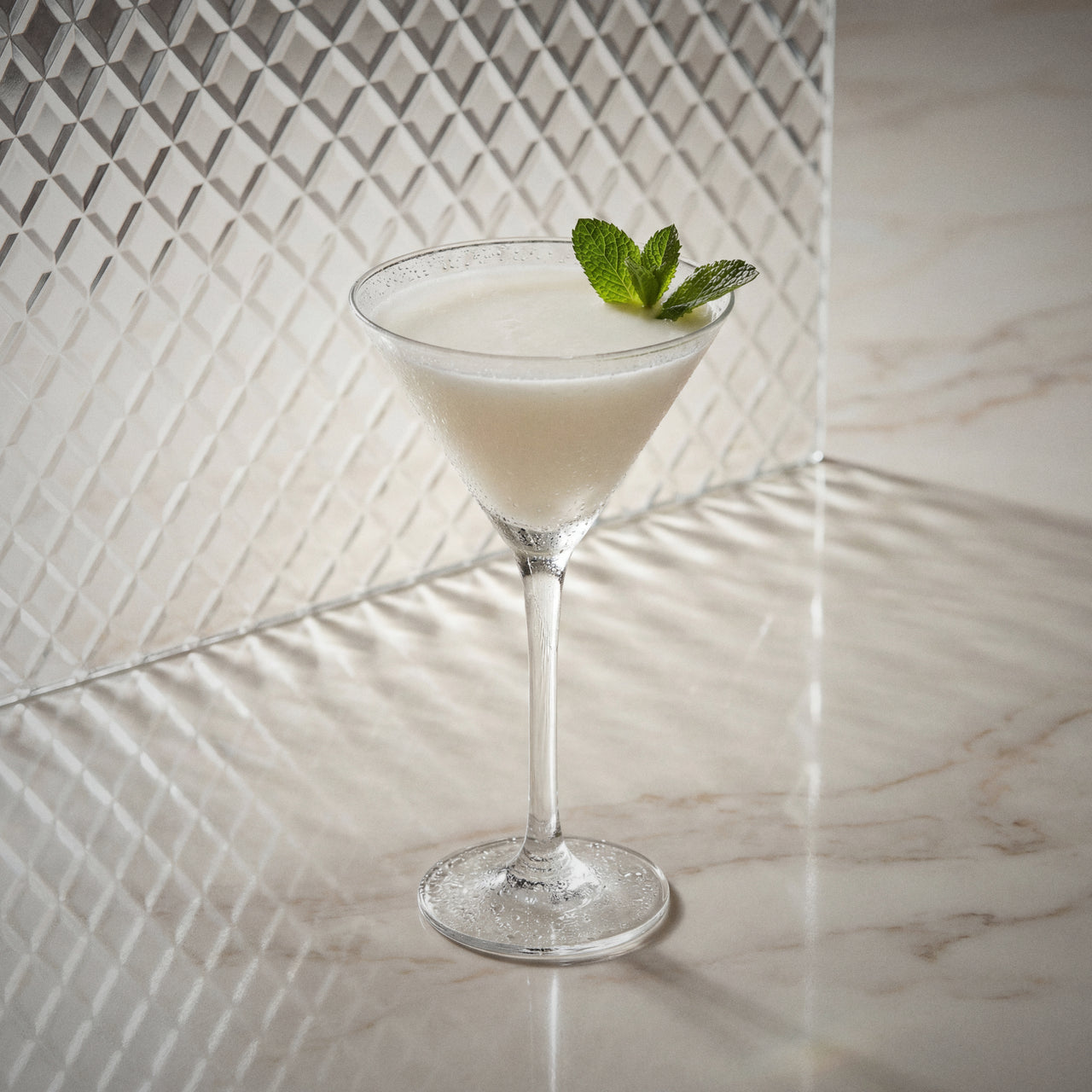 albino grasshopper cocktail with natural yoghurt liqueur, peppermint white liqueur, and cacao white liqueur in a martini glass with a mint leaf on a marble surface.