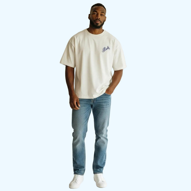 A man wearing a whi-colored t-shirt with a small blue graphic on the left chest, paired with blue jeans and white sneakers