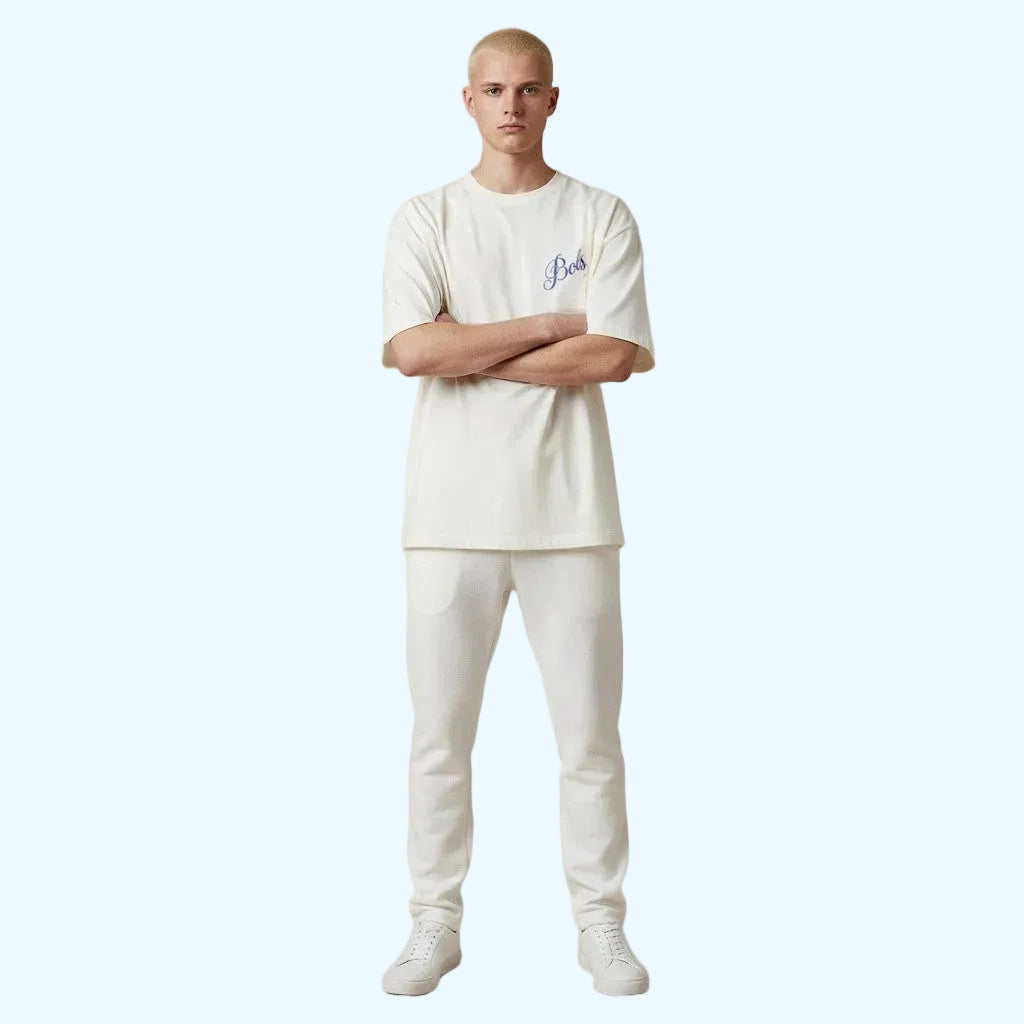 A man stands with arms crossed wearing a white crewneck t-shirt with a small blue Bols logo on the left chest, matching white pants, and white sneakers.
