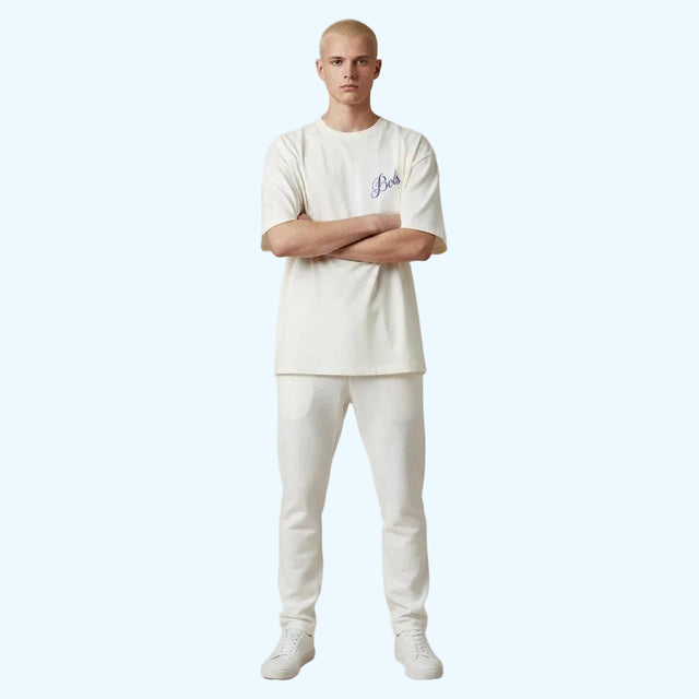 A man stands with arms crossed wearing a white crewneck t-shirt with a small blue Bols logo on the left chest, matching white pants, and white sneakers.