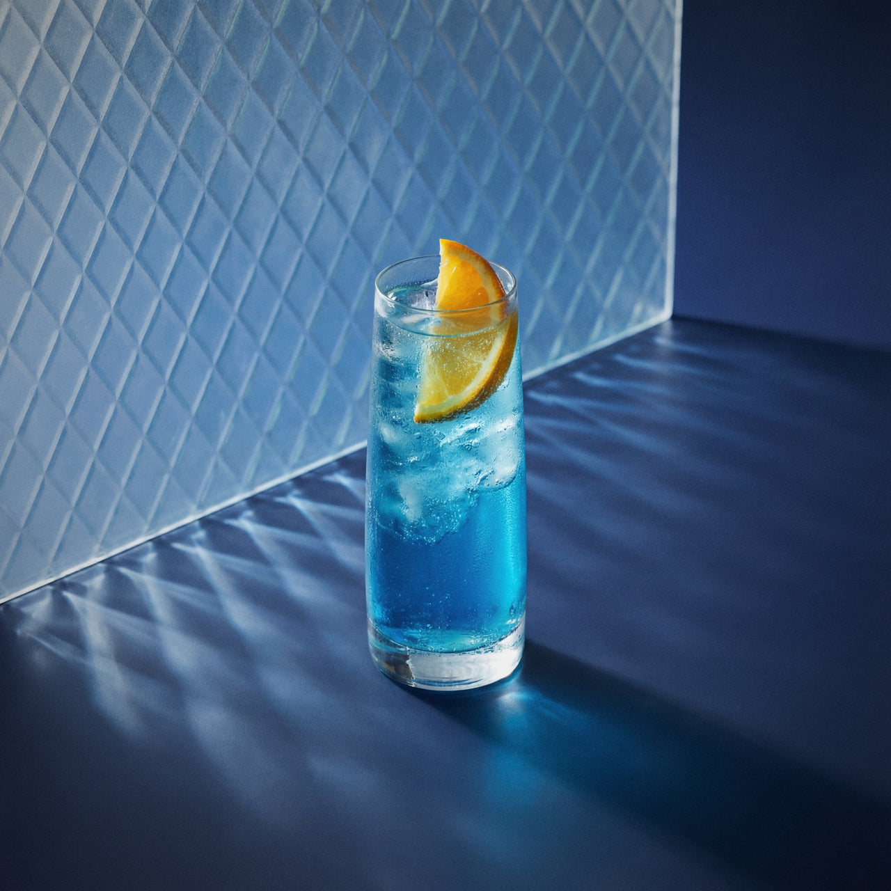 blue buck cocktail with blue liqueur in a highball glass with an orange wedge  against a blue textured wall.