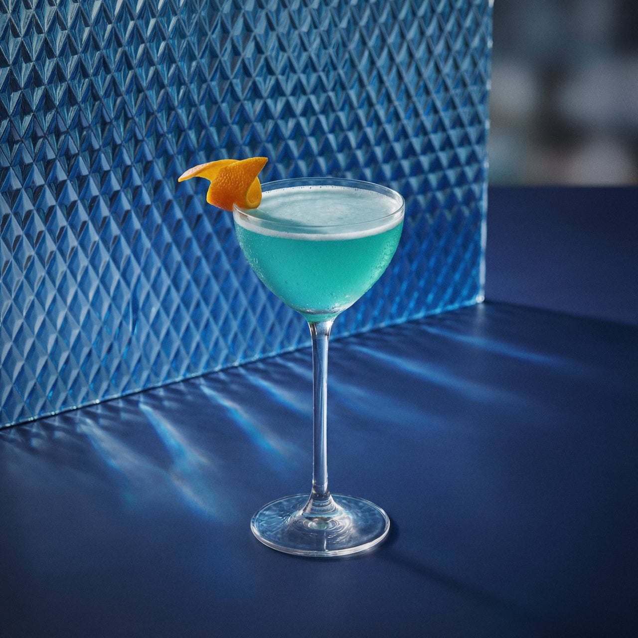 blue martini cocktail with blue liqueur and vodka in a nick & nora glass with an orange peel on a textured surface
