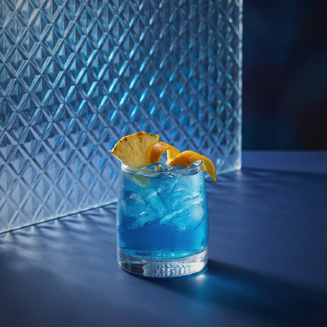 blue bird cocktail with blue liqueur in a rocks glass with a pineapple wedge and orange twist on a textured surface