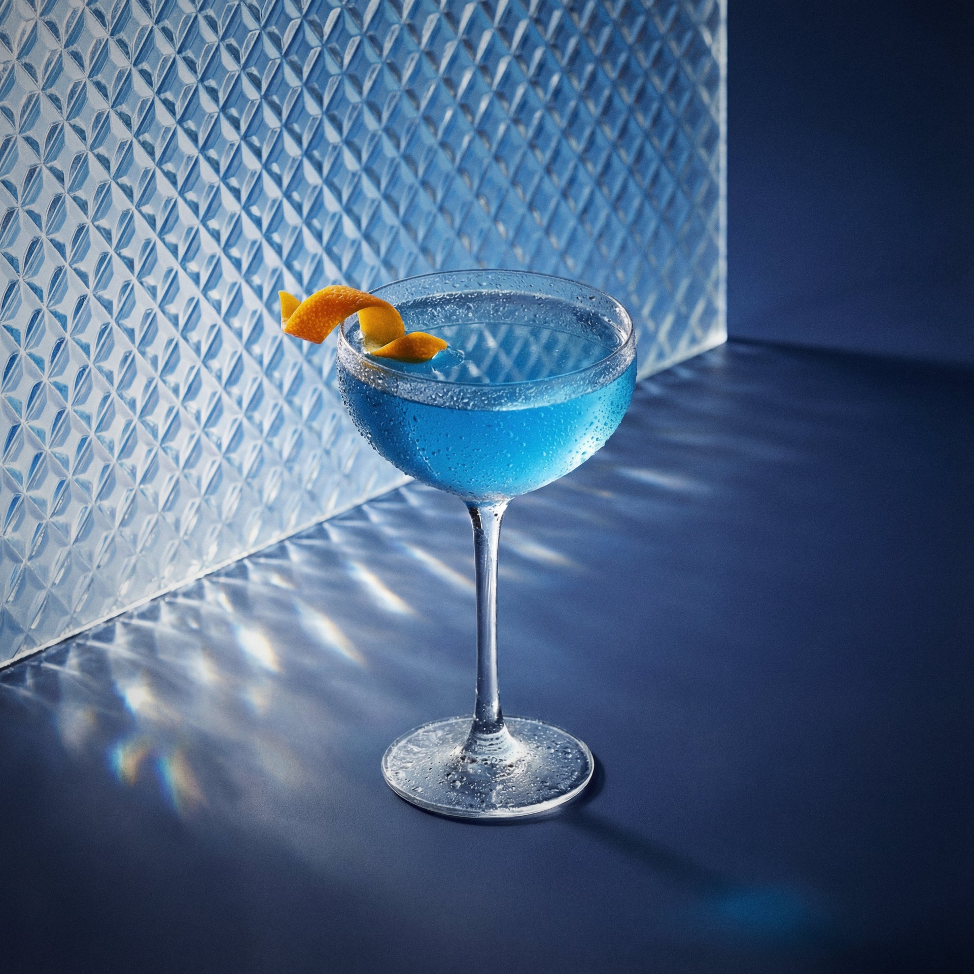 Blue pegu cocktail with blue liqueur and gin in a coupe glass with an orange twist against a textured blue wall.