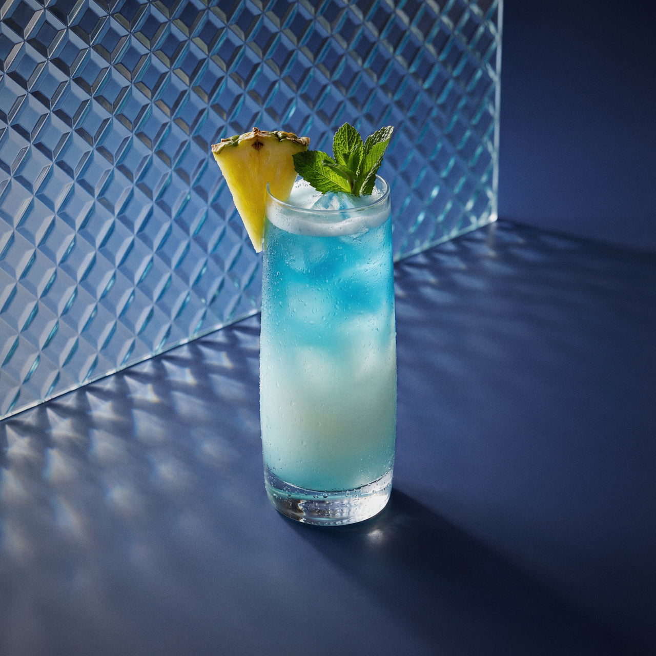 blue swimming pool cocktail with blue liqueur and coconut liqueur in a highball glass with a pineapple wedge and mint sprig on a textured surface