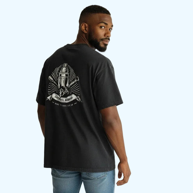A man wearing a black graphic t-shirt with a bols cocktail academy print on the back, paired with blue jeans, photographed against a light gray studio background.