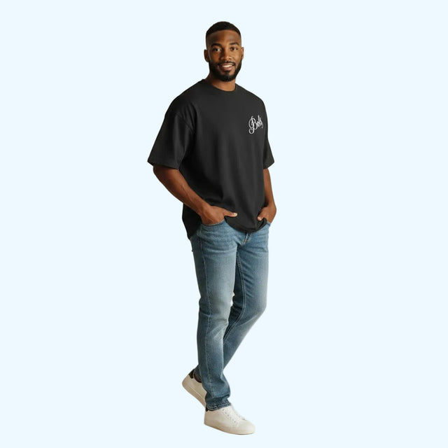 A man modeling a black crewneck t-shirt with a small white graphic on the left chest, paired with blue jeans and white sneakers, standing against a solid beige studio background.