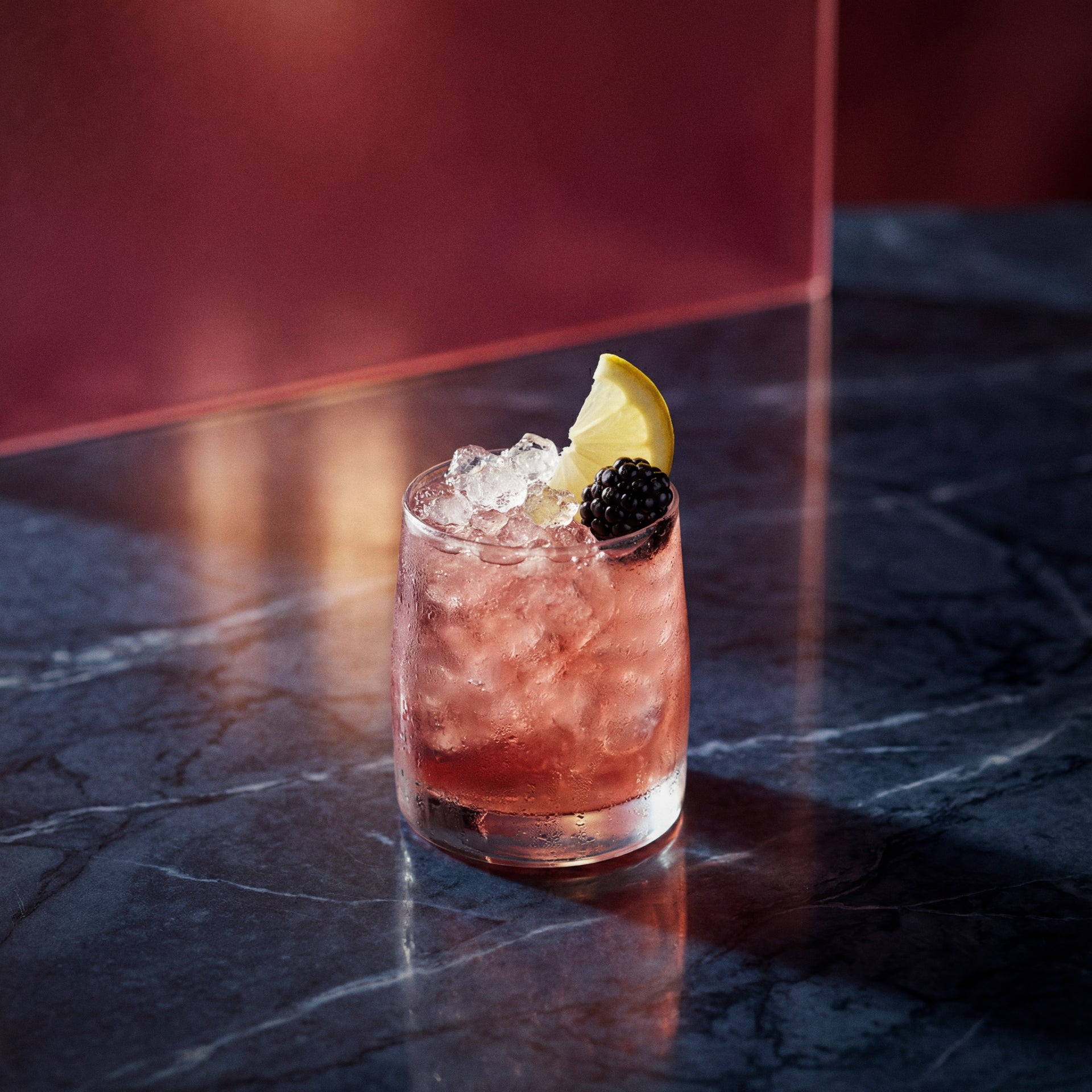 bramble cocktail with creme de cassis liqueur and gin in a rocks glass with a blackberry and lemon wedge on a textured surface
