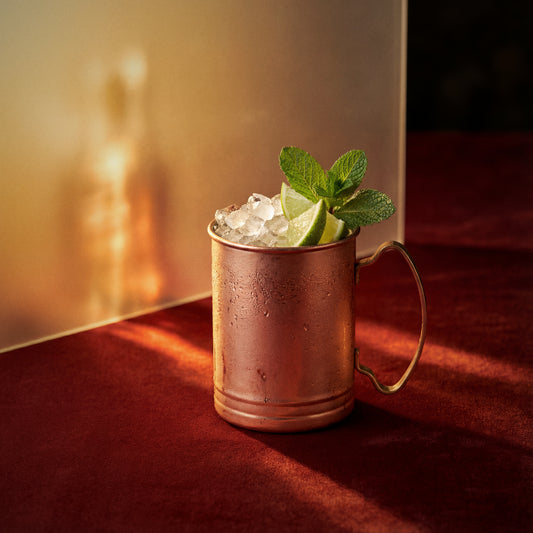 dry orange mule cocktail with dry orange liqueur in a copper mug garnished with lime and mint leaves on a red surface