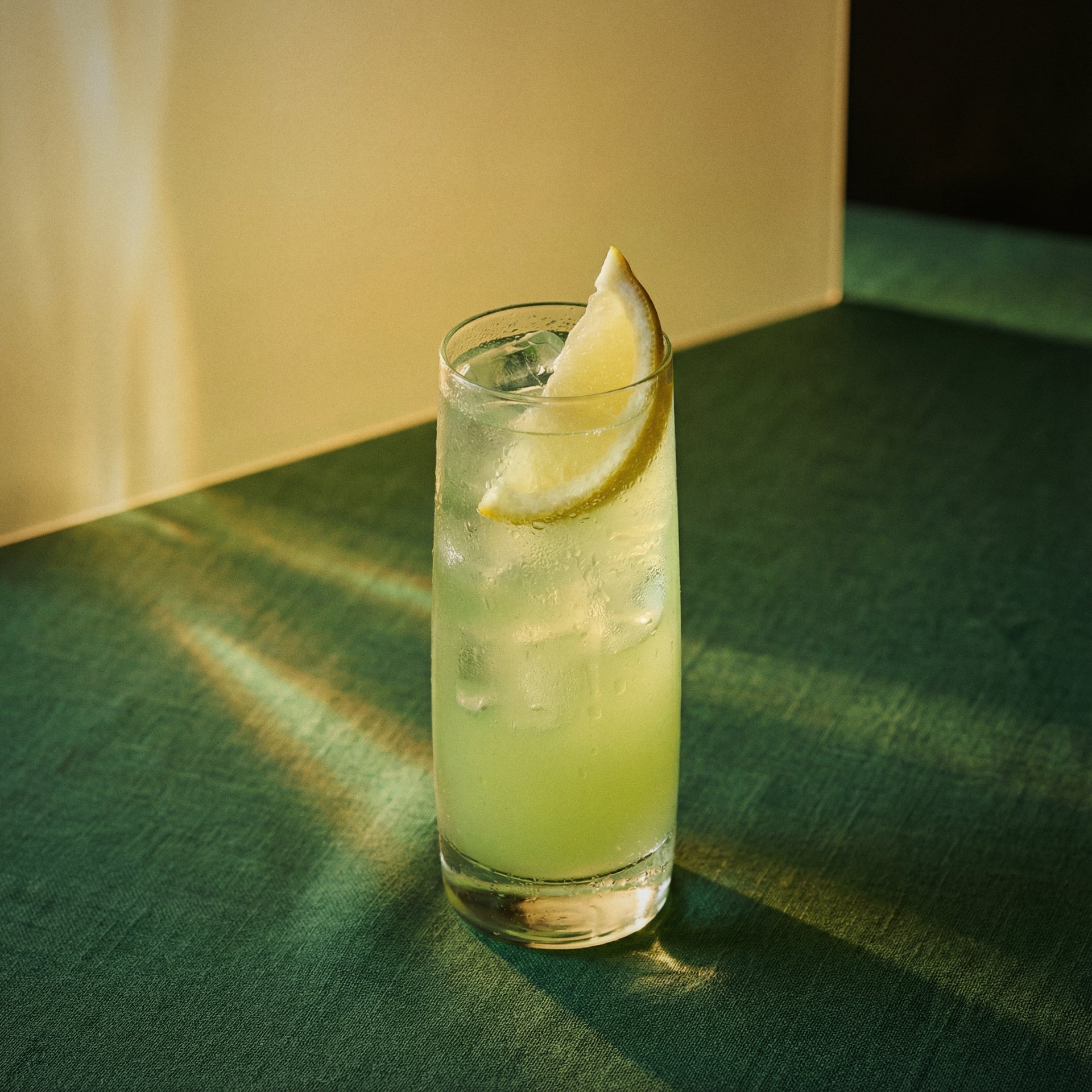 green banana collins cocktail with green banana liqueur and genever in a highball glass with a lemon wedge on a green surface