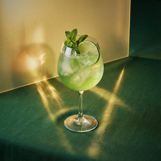 Green banana spritz cocktail with green banana liqueur and lime and mint in a large wine glass on a green surface