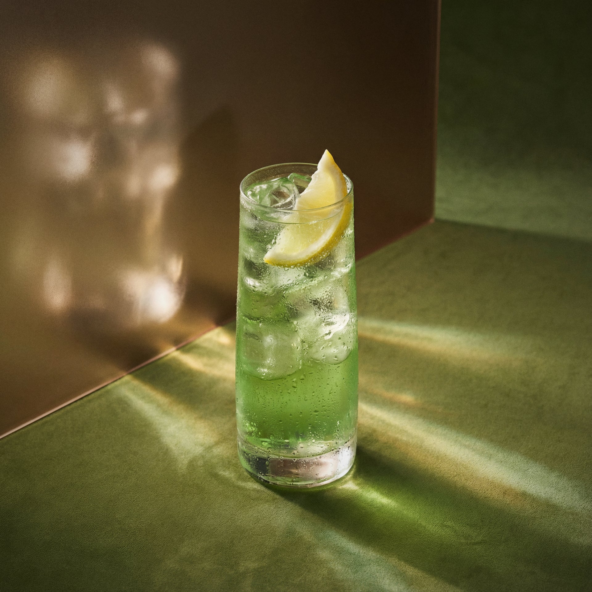 kiwi collins cocktail with lemon wedge in a highball glass made with kiwi liqueur and gin on a reflective surface