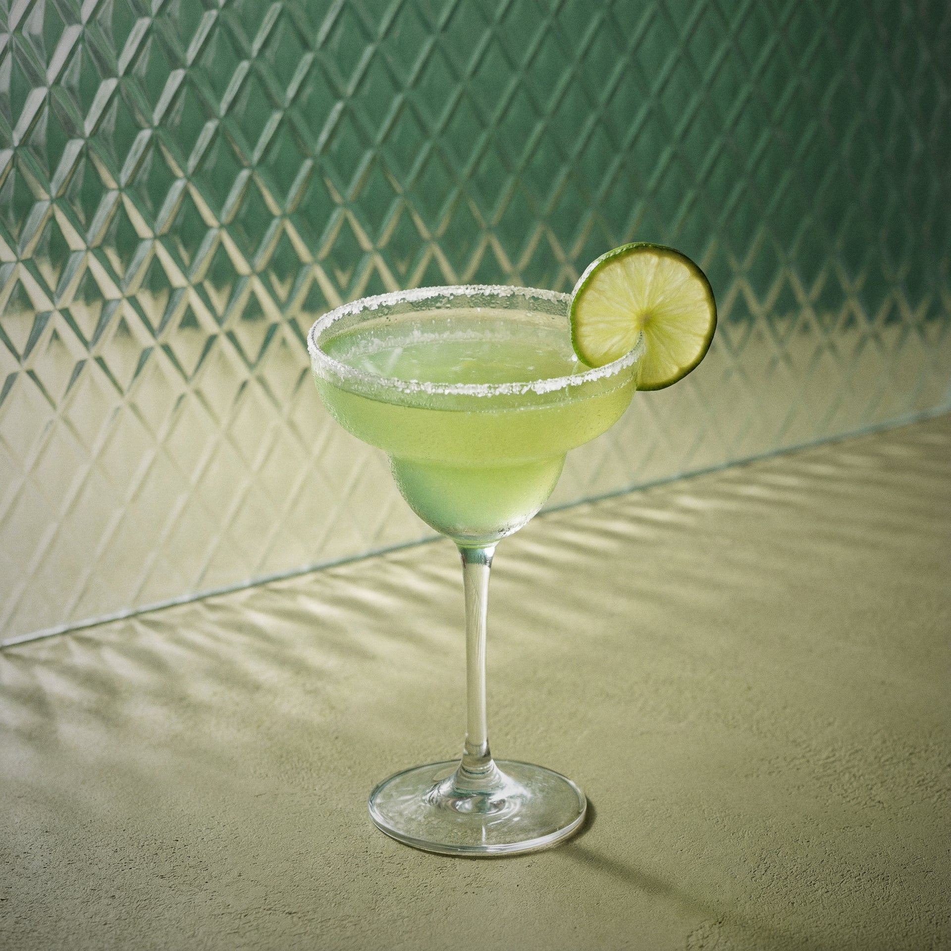 melon margarita cocktail with melon liqueur and triple sec liqueur in a margarita glass and a lime wheel in a green textured surface