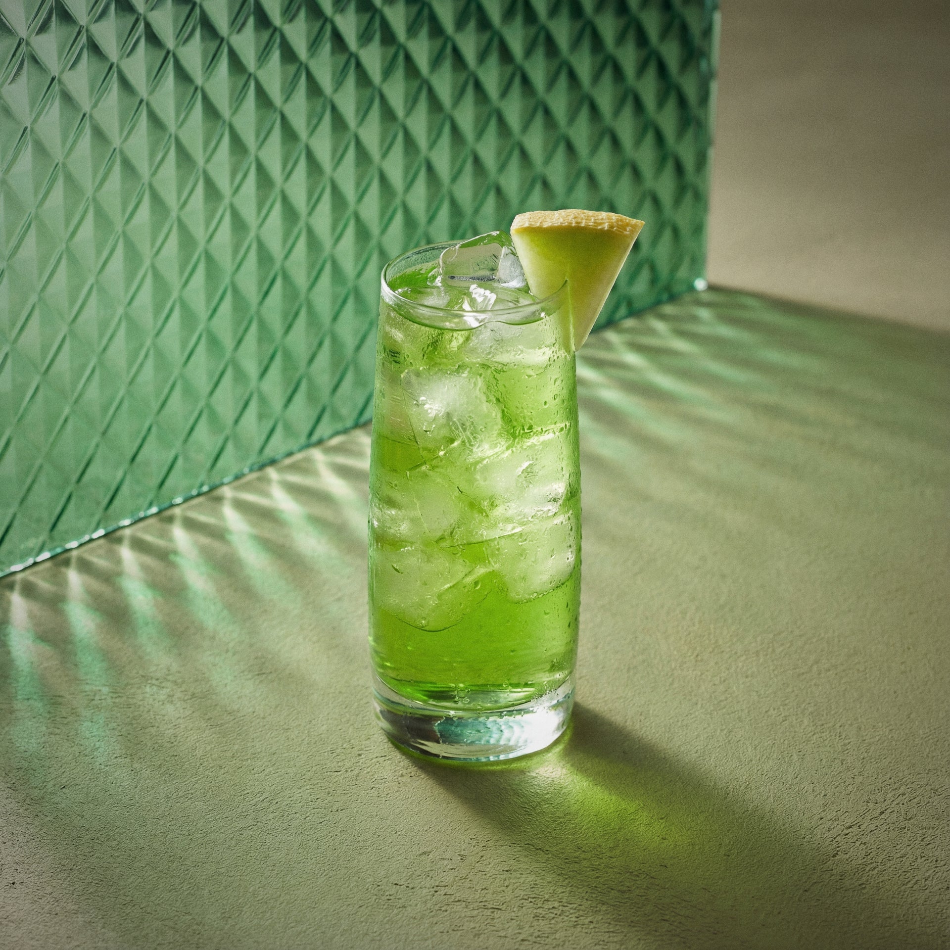 melon tonic cocktail with melon liqueur and a lime wedge in a glass against a textured green background