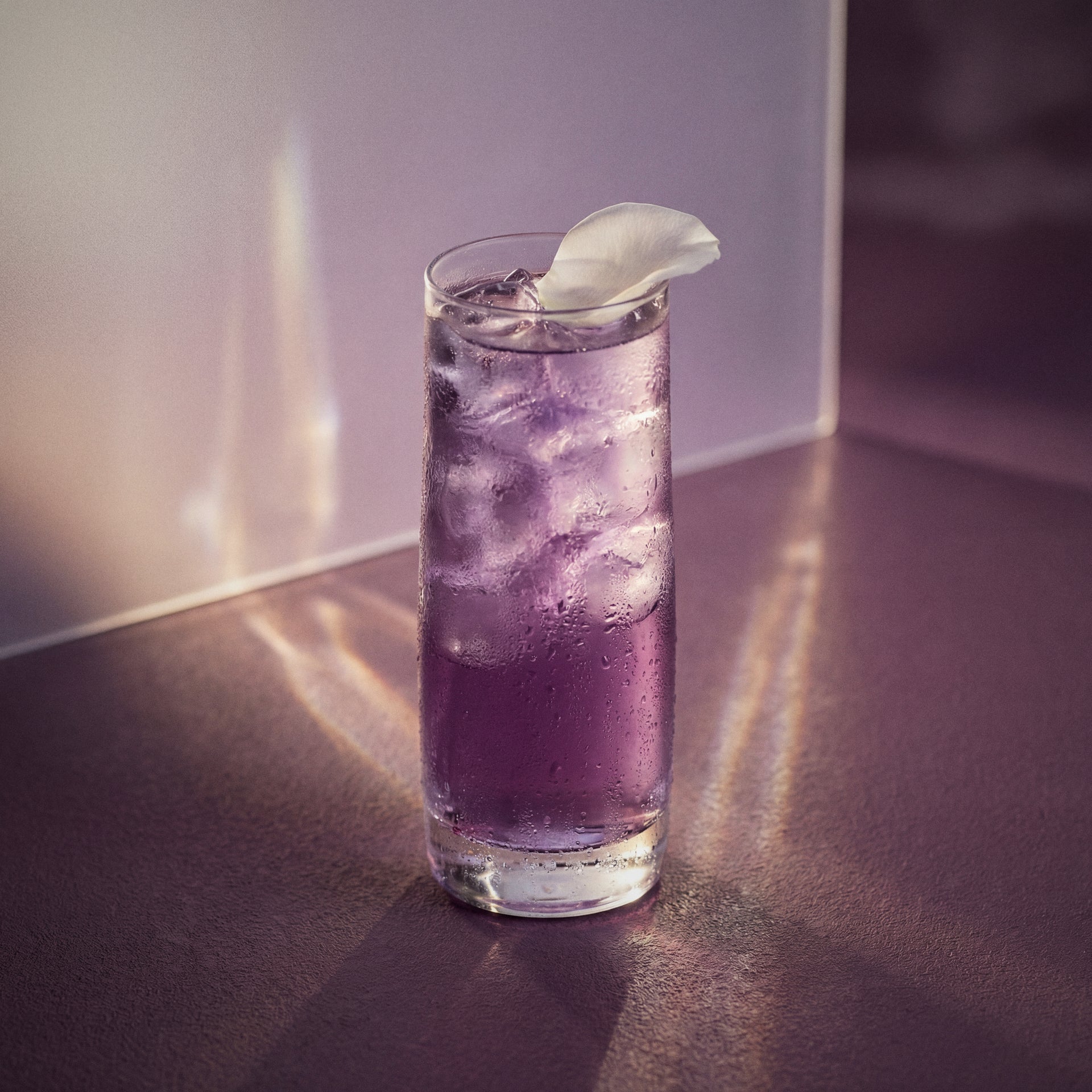 purple parfait amour cocktail with parfait amour liqueur with ice and a petal in a glass on a reflective surface.
