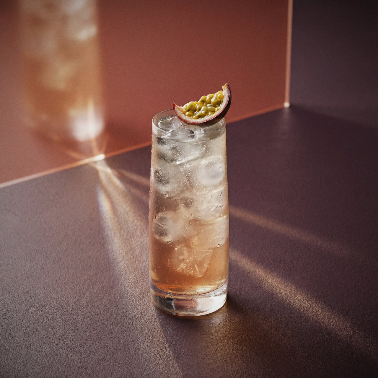 passionfruit collins cocktail with passionfruit liqueur and barrel aged genever with ice and a slice of passionfruit on a reflective surface