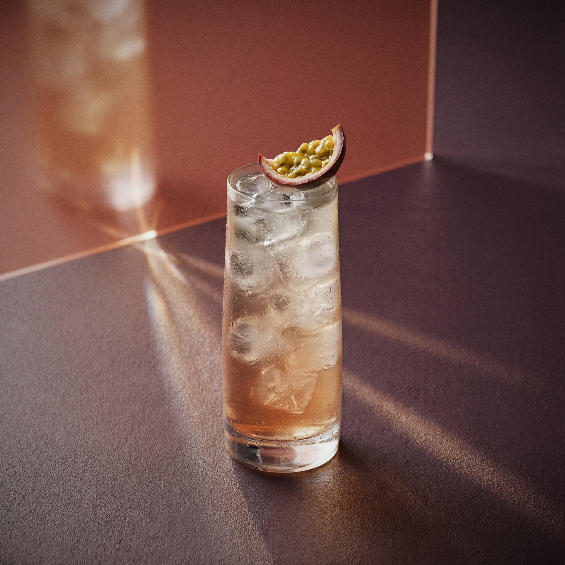 passionfruit collins cocktail with passionfruit liqueur and barrel aged genever with ice and a slice of passionfruit on a reflective surface