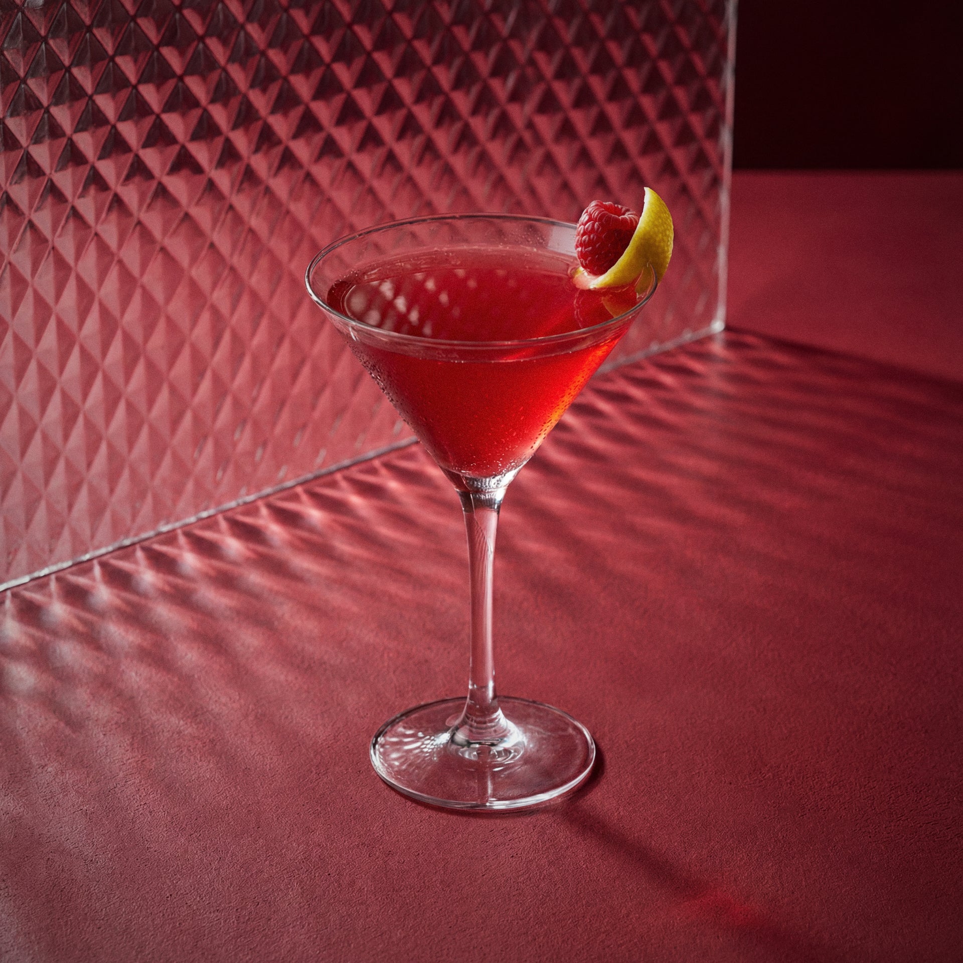 raspberry martini cocktail with raspberry liqueur and vodka in a martini glass with a lemon twist on a red textured surface