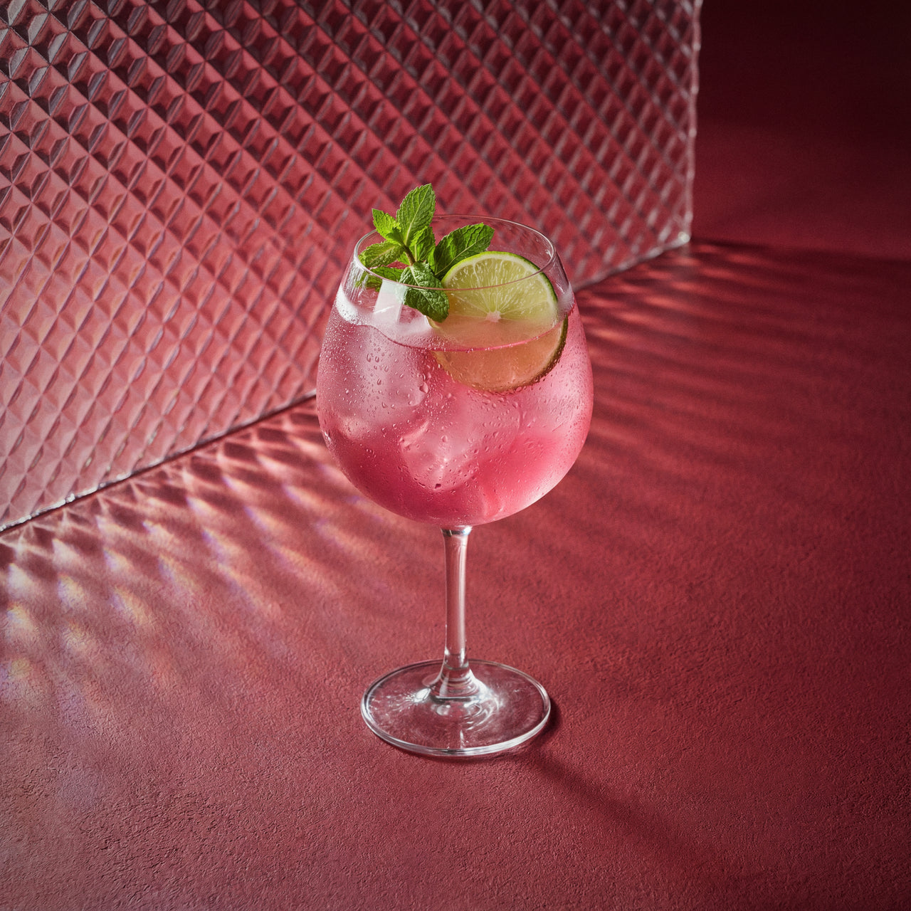 raspberry spritz cocktail with lime and mint in a coppa glass on a red textured surface 