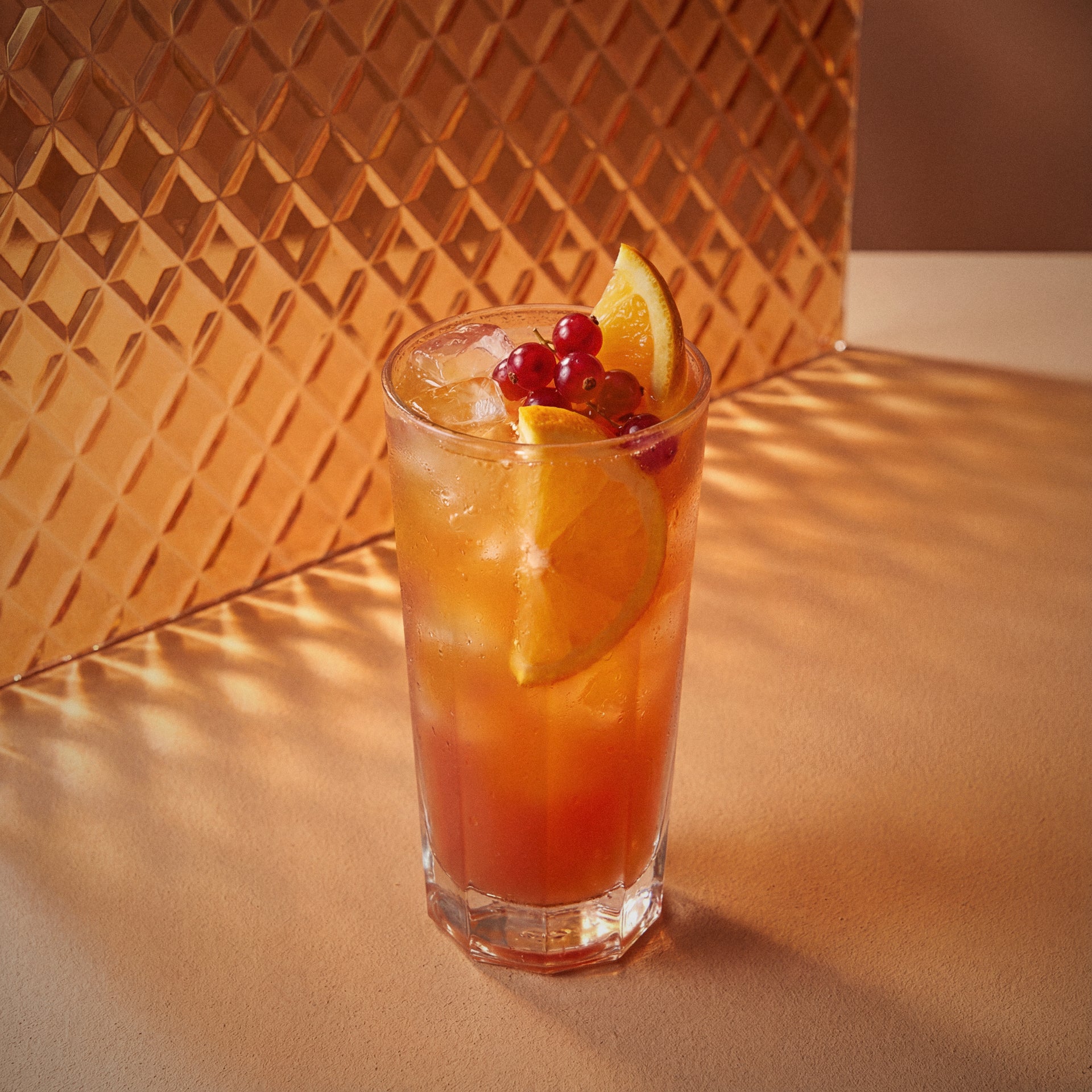 sex on the beach cocktail with peach liqueur and vodka, and orange slices and berries in a glass against an orange textured wall.