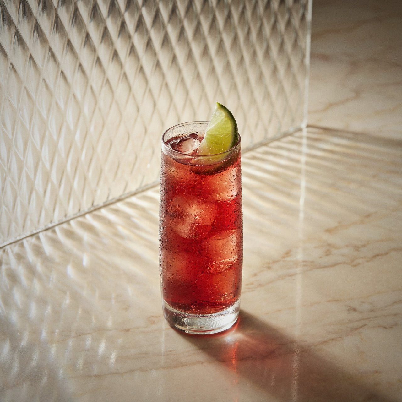 vanilla berry cooler cocktail with ice and a lime wedge and strawberry liqueur and vanilla liqueur in a highball glass on a marble surface.