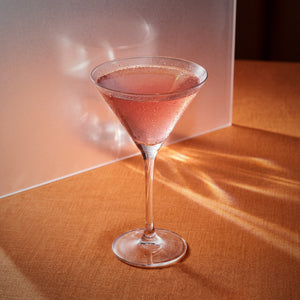Verano cocktail in a martini glass with pink grapefruit liqueur on a wooden surface with sunlight casting shadows